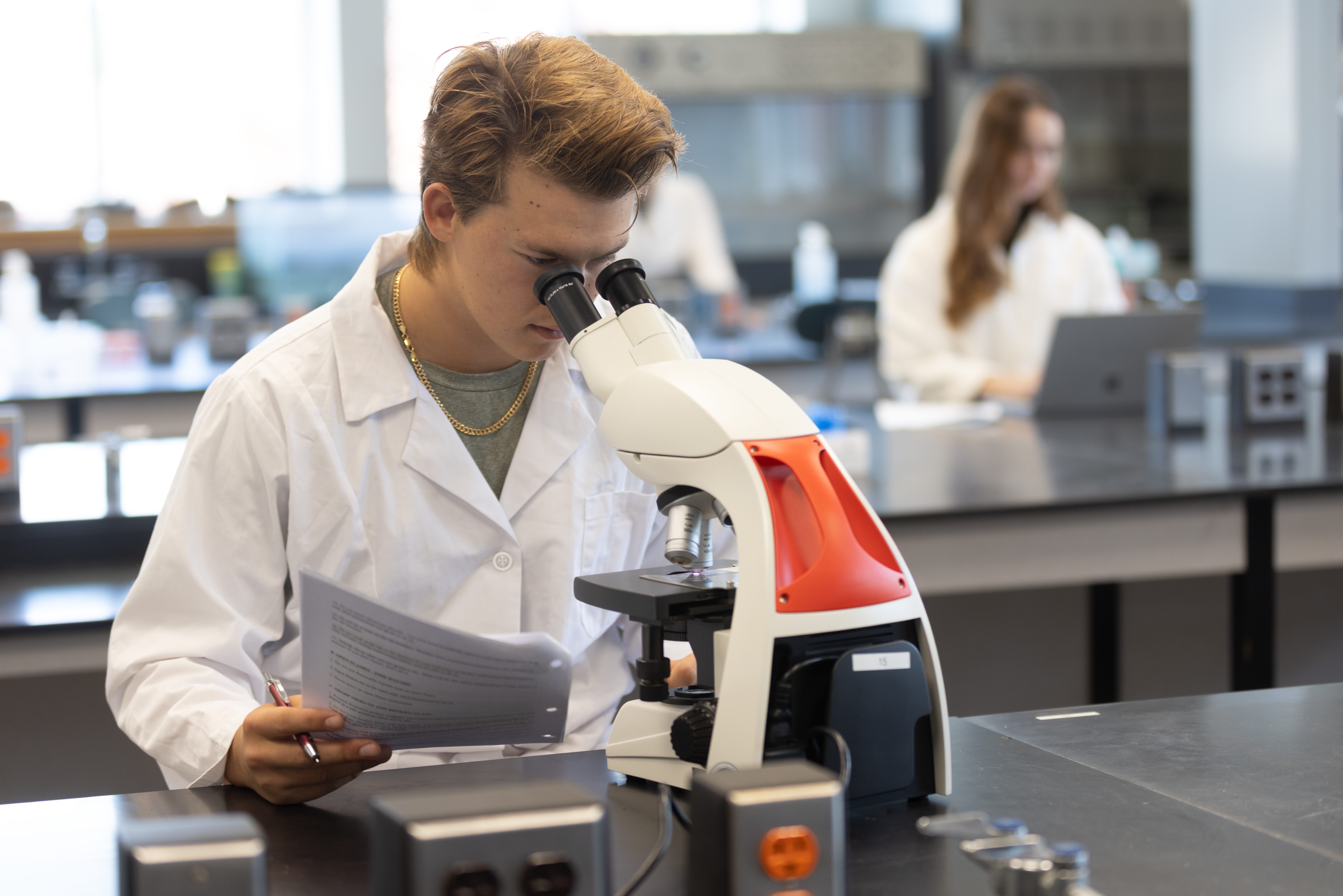 A male student in the Biology lab looking into a microscope with two female students in the background.