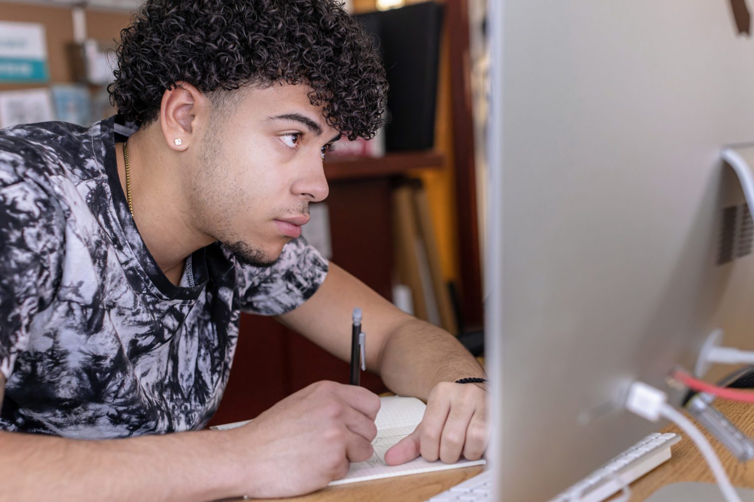 A male student sitting at a desk in front of a computer and writing.