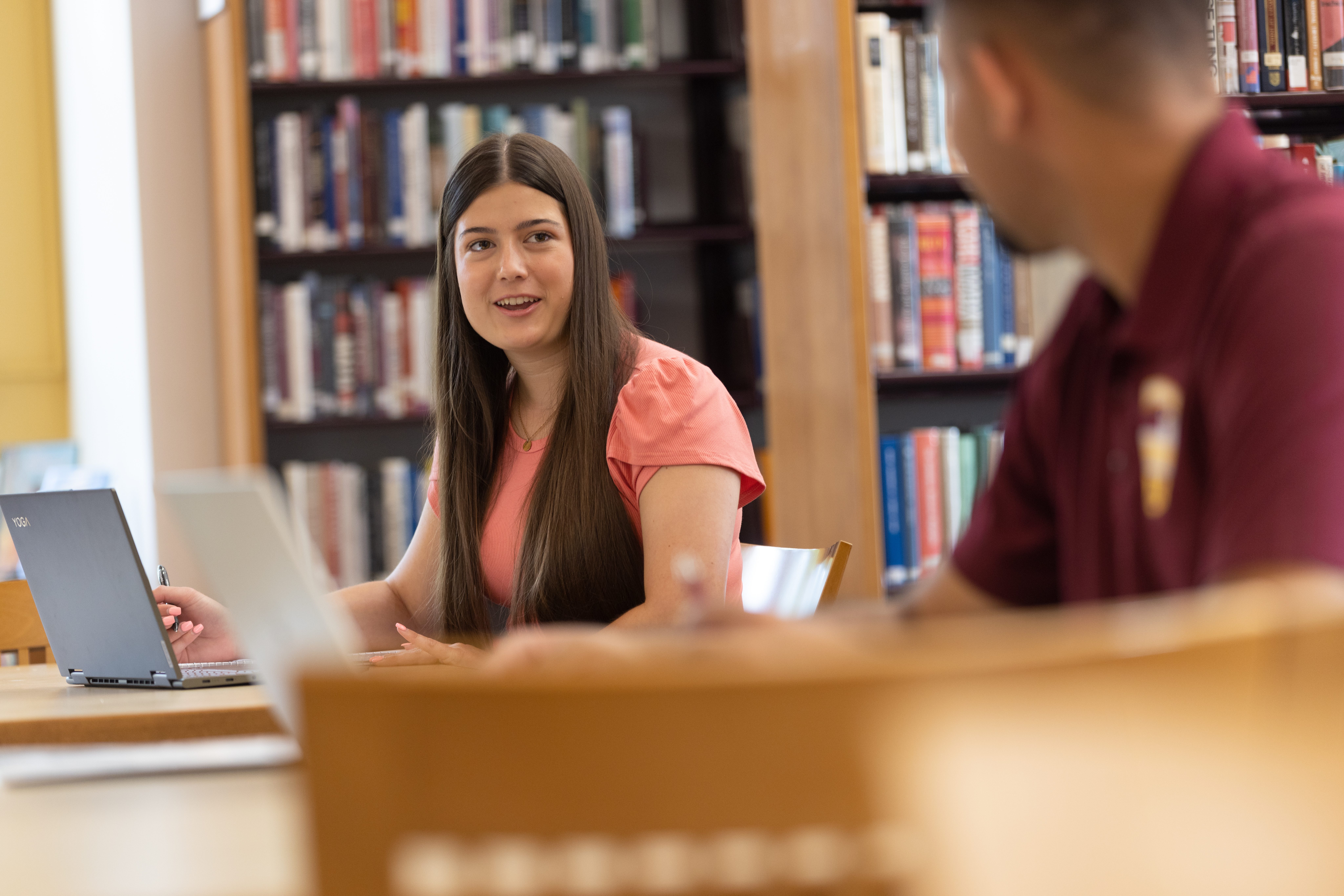 A male and female student sitting at tables in library with their laptops in front of them and talking