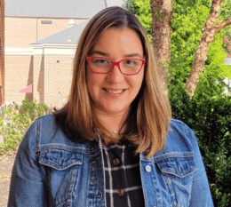 Nydia smiling with greenery and brick building in background wearing glasses blue shirt and jean jacket