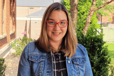 Nydia smiling with greenery and brick building in background wearing glasses blue shirt and jean jacket