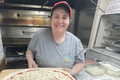 Victoria smiling at work wearing tshirt and hat holding a pizza ready to cook in the oven