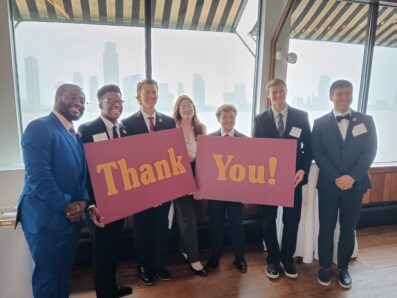 7 students standing at President's Club dinner holding thank you sign and smiling