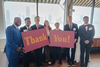 7 students standing at President's Club dinner holding thank you sign and smiling