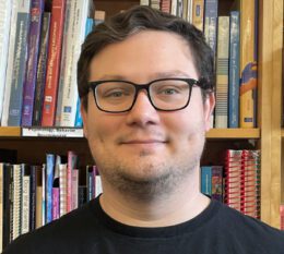Andrew smiling wearing glasses and black shirt with book stacks in background