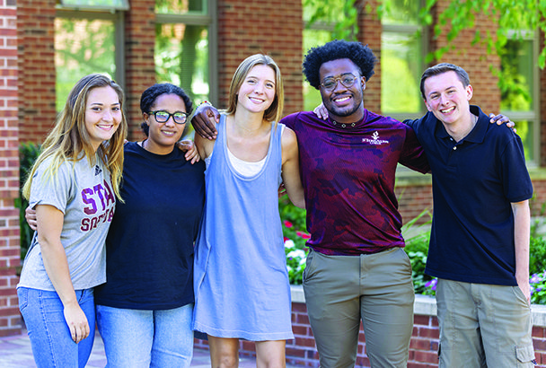Group of five students in front of building entrance.